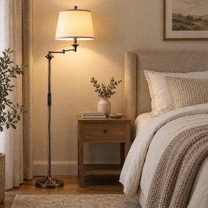 May include: A bronze floor lamp with an adjustable swing arm and a white fabric drum shade. The lamp features a slender metal pole, a circular base, and a warm light emitting from the shade, positioned next to a wooden nightstand.