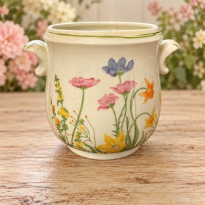 May include: A cream-colored ceramic pot with two side handles, featuring a painted botanical design of pink, yellow, and blue wildflowers. The rim is accented with a thin green line, and the exterior surface displays a variety of floral stems.