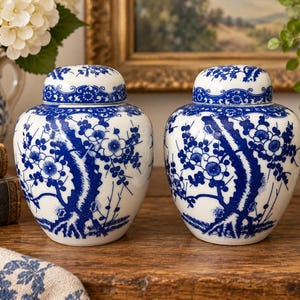May include: A pair of white ceramic ginger jars with lids, each featuring a hand-painted blue floral pattern depicting blossoming tree branches. The jars have a rounded body and a matching removable lid with a decorative blue border design.