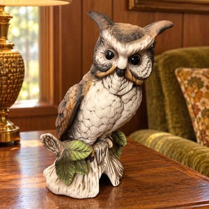 May include: A ceramic owl figurine perched on a textured tree stump base. The owl features detailed gray, brown, and white plumage with large black eyes. The base includes sculpted green leaves and a light-colored bark texture on the stump.