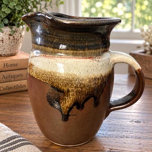 May include: A ceramic pitcher featuring a multi-toned glaze with a dark brown base, a cream-colored middle section, and a dark brown rim. The glaze exhibits a drip effect where the darker colors run down into the lighter cream-colored center.
