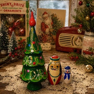 May include: A green wooden Christmas tree ornament with a red pointed top, gold glitter accents, and colorful dots. Beside it stands a wooden Santa Claus figurine and a small white snowman with a blue hat and scarf, all arranged on a lace doily.