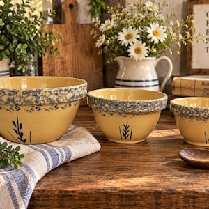 May include: A set of three yellow ceramic mixing bowls of varying sizes, each featuring a blue speckled rim and a simple blue sprig design on the front. The bowls are arranged in a row, showcasing a consistent color palette and decorative pattern.