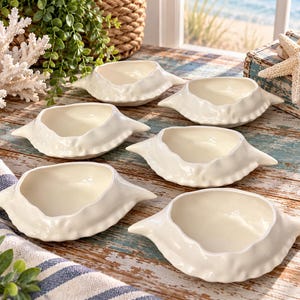 May include: A set of six off-white ceramic bowls, each shaped like a crab shell with a textured, organic rim and a smooth, glossy interior. These small, shallow dishes feature pointed side extensions and a uniform, neutral cream-colored finish.
