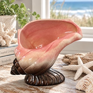 May include: A ceramic conch shell vessel featuring a gradient color scheme that transitions from a deep dark brown at the base to a soft cream in the middle and a vibrant pink hue on the interior and upper rim, designed with a glossy finish.