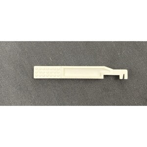 May include: A light beige plastic tool featuring a rectangular handle with a textured grid pattern of small raised squares on one end and a flat, elongated body that tapers into a notched, hook-like mechanism on the opposite end.