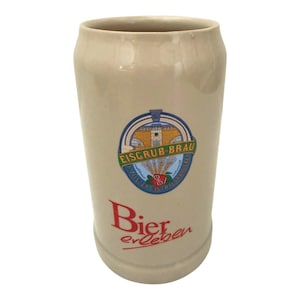 May include: A beige ceramic beer mug with a smooth finish. The front features a circular logo with the text Eisgrub-Brau and a building illustration. Below the logo, the red text Bier erleben is printed in a stylized font on the mug surface.
