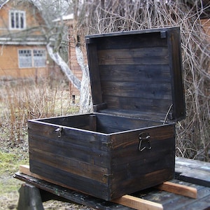 Op de afbeelding: Een rechthoekige houten opbergkist met een donkere, gebrande afwerking en een opengeklapt deksel. De kist heeft een zichtbare houtnerf, metalen handgrepen aan de zijkanten en een sluiting aan de voorzijde, opgebouwd uit horizontale houten planken.