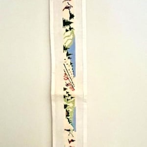 Pode incluir: Uma tira de tecido branco longa e estreita com um padrão bordado repetitivo de esquiadores, árvores perenes e picos de montanhas azuis. O design é orientado verticalmente e inclui pequenas figuras em fios de cores vermelha, azul e preta.