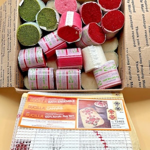 May include: A cardboard box filled with bundles of pre-cut acrylic rug yarn in shades of green, pink, red, and cream, alongside a Bucilla Bath Ensemble canvas kit labeled Hearts Delight No. 12403, featuring a grid pattern with red stitched accents.