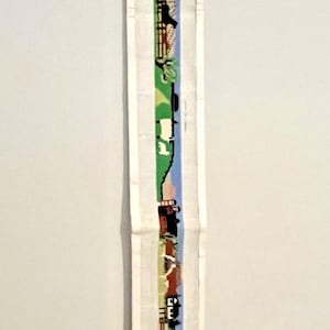 May include: A long, narrow white fabric strip featuring a vertical embroidered scene of a rural landscape. The design includes green fields, brown fences, small houses, and various animals depicted in a repeating pattern along the center length.