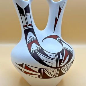 Puede incluir: Un jarrón de boda de cerámica blanca con un asa central y dos bocas. La superficie está decorada con patrones geométricos y motivos de aves estilizadas en pintura negra y roja oscura, creando un diseño simétrico en el cuerpo redondeado.