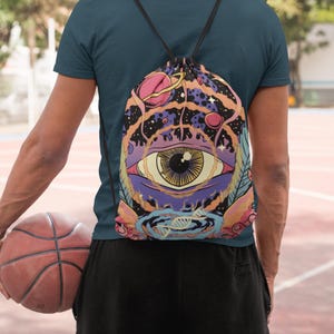 May include: A drawstring backpack featuring a vibrant, surreal graphic print of a large central eye surrounded by swirling cosmic elements, planets, stars, and a DNA helix structure in shades of purple, orange, pink, and black with black cords.