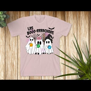 May include: A light pink t-shirt featuring a graphic of three white ghosts holding small jars. The text "LAS BOOO-RRRACHAAS" is printed above the ghosts, accompanied by illustrations of spiderwebs, bats, a broom, and a witch hat.