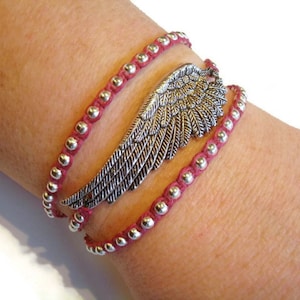 May include: A red beaded wrap bracelet with a silver wing charm. The bracelet is layered and has a delicate, feminine look.