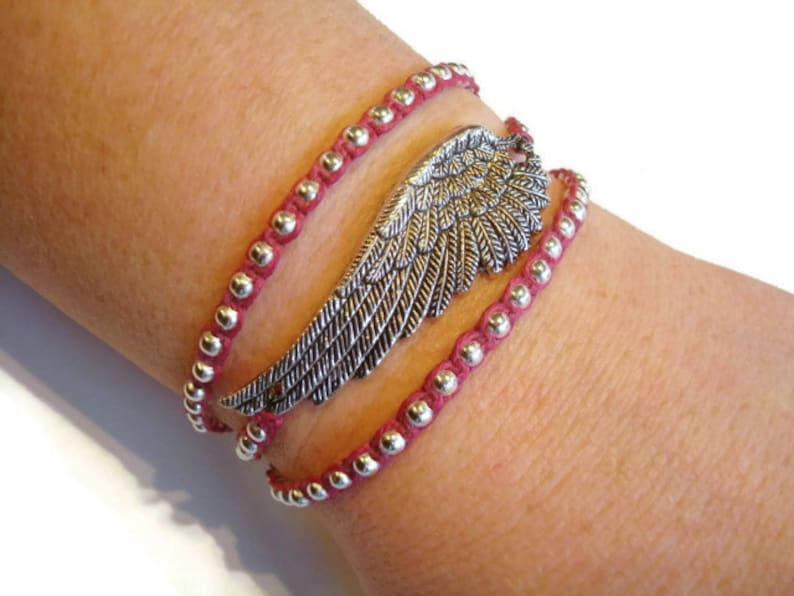 May include: A red beaded wrap bracelet with a silver wing charm. The bracelet is layered and has a delicate, feminine look.