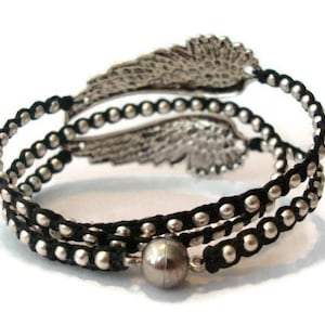 K&ouml;nnte beinhalten: Ein schwarzes und silbernes Armband mit einem silbernen Fl&uuml;gel-Anh&auml;nger. Das Armband besteht aus schwarzem Band und silbernen Perlen.