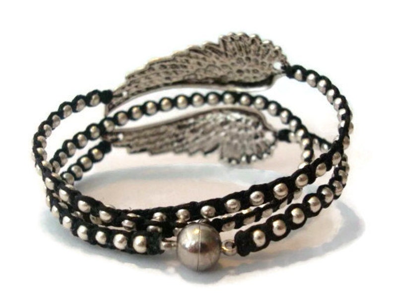 K&ouml;nnte beinhalten: Ein schwarzes und silbernes Armband mit einem silbernen Fl&uuml;gel-Anh&auml;nger. Das Armband besteht aus schwarzem Band und silbernen Perlen.