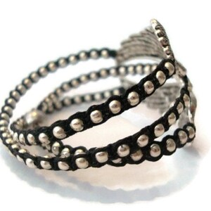 K&ouml;nnte beinhalten: Ein schwarzes und silbernes Perlenarmband mit einem dekorativen Metalldesign. Das Armband besteht aus mehreren Str&auml;ngen schwarzer Schnur mit silbernen Perlen.