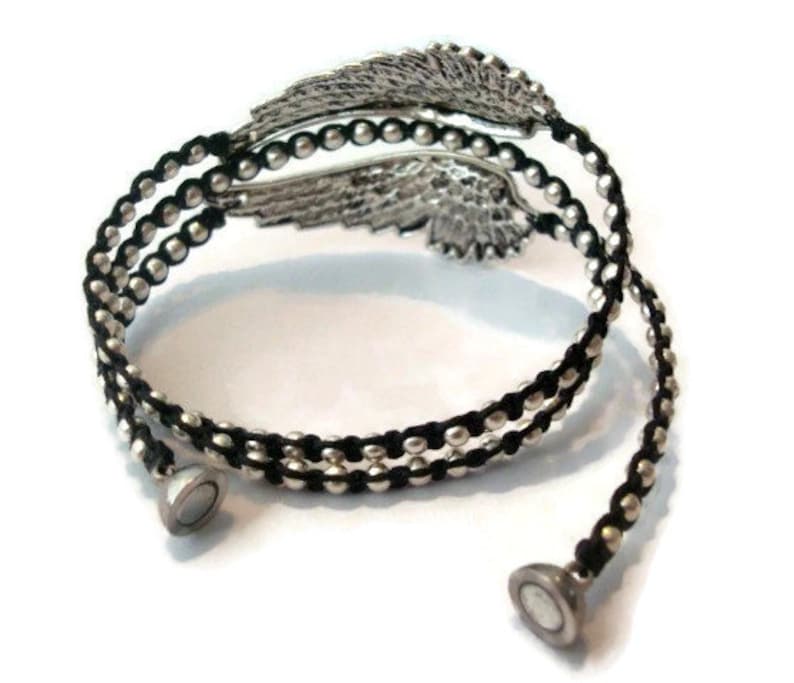 K&ouml;nnte beinhalten: Ein schwarzes und silbernes Wickelarmband mit einem silbernen Fl&uuml;gel-Anh&auml;nger. Das Armband besteht aus schwarzem Band und silbernen Perlen. Das Armband hat einen magnetischen Verschluss.