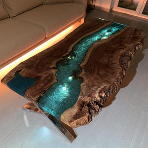 May include: A rectangular coffee table featuring a live edge wood slab with a central river-like inlay of translucent turquoise resin. The resin section is illuminated from within by small lights, and the table rests on a polished metal X-shaped base.