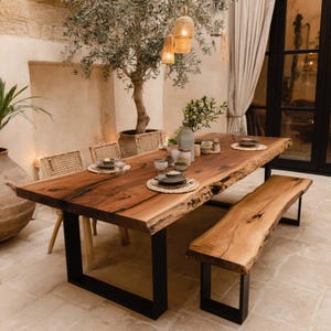Peut inclure: Une table à manger en bois à bord brut avec un banc assorti, présentant des motifs de grain de bois naturel et des pieds en U en métal noir. La table est disposée avec des sets de table tressés, des bols en céramique et des petits vases.