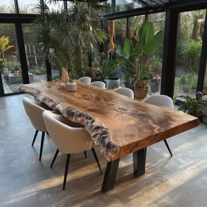 Peut inclure: Une grande table de salle à manger en bois à bord brut avec un grain naturel et des bords en écorce, soutenue par deux pieds en métal noir. La table est entourée de plusieurs chaises rembourrées blanc cassé au tissu texturé.
