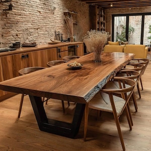 Puede incluir: Una gran mesa de comedor rectangular con un tablero de madera maciza de borde natural y una base de metal oscuro en forma de U. La mesa está rodeada por varias sillas de madera con asientos trenzados. Un jarrón de cristal con flores secas está sobre la mesa.