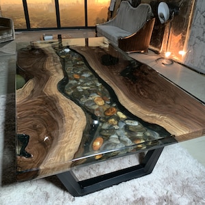 May include: A rectangular dining table featuring two large slabs of natural dark brown wood with a prominent grain pattern, separated by a central river-like inlay of clear resin filled with smooth, multi-colored river stones and black metal legs.