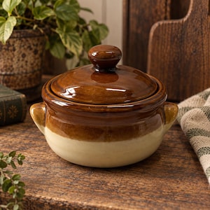 Pode incluir: Uma panela de cerâmica redonda com tampa, apresentando um design de dois tons com uma seção superior esmaltada em marrom escuro e uma base na cor creme. A panela inclui duas pequenas alças laterais e um botão central na tampa.