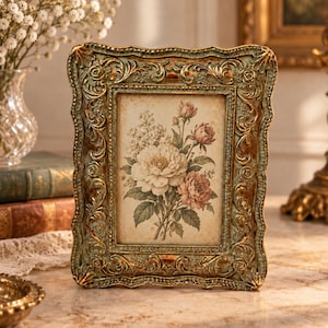 May include: A rectangular picture frame with an ornate, scalloped border finished in a distressed sage green and metallic gold. The frame contains a print of a floral bouquet featuring white and pink roses with green leaves on a cream background.