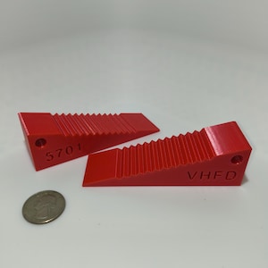 May include: Two bright red door wedges with a serrated top surface. One wedge is embossed with the number 5701 and the other with the letters VHFD. Each wedge features a circular hole at the taller end. A US quarter coin is placed nearby for scale.