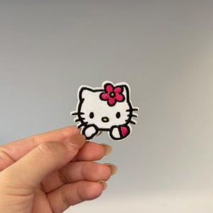 May include: An embroidered patch featuring the white face of a cartoon cat with black outlines, three whiskers on each side, a small yellow nose, and a bright pink flower with a dark center positioned above the right ear of the character.