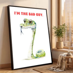 Könnte beinhalten: Ein gerahmter Kunstdruck mit einer Aquarellillustration einer grünen Schlange mit großen Augen und einer blauen gespaltenen Zunge. Der Text "I'M THE BAD GUY." ist in roten Großbuchstaben über der Schlange auf weißem Hintergrund gedruckt.