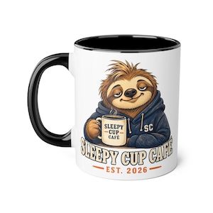 May include: A white ceramic mug with a black handle and interior rim. The front features a graphic of a brown sloth wearing a dark blue hoodie, holding a smaller mug labelled SLEEPY CUP CAFE. Below the sloth, the text reads SLEEPY CUP CAFE EST. 2026.
