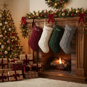 Può includere: Quattro calze natalizie in maglia a trecce color bordeaux, bianco, verde scuro e grigio appese a una mensola in legno. Ogni calza presenta un risvolto con i nomi ricamati Charlie, Sophie, Liam e Red in un carattere corsivo.