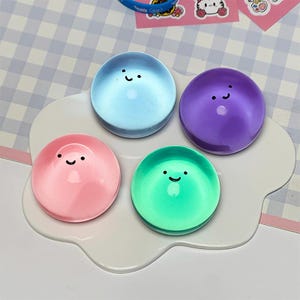 May include: Four circular, smooth, glossy objects in light blue, purple, pink, and green, each featuring a simple black printed smiley face. These items are arranged on a white, cloud-shaped base, creating a set of colorful, rounded decorative pieces.