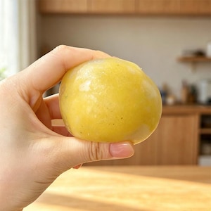 May include: A round, smooth, yellow-toned food item with a slightly translucent, gelatinous texture. The object has a soft, pliable appearance with subtle variations in color, featuring faint speckles throughout its surface.