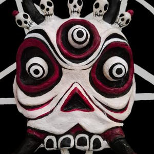 May include: A three-eyed mask featuring a white base with black and deep red accents. The mask displays a central eye, two large side eyes, a triangular mouth, and four small skulls along the top edge, with two curved horns extending from the sides.