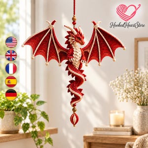 Op de afbeelding: Een hangend gehaakt drakenornament met een dieprood lichaam en crèmekleurige accenten op de vleugels, borst en staart. De draak heeft gedetailleerde steken, kleine hoorns en hangt aan een koord met decoratieve kralen aan de boven- en onderkant.