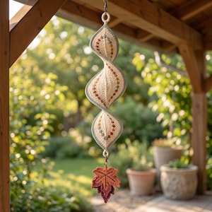 Puede incluir: Un móvil decorativo de ganchillo en forma de espiral, hecho de tela color crema con bordes verde salvia y una fila vertical de pequeñas cuentas multicolores. Una hoja de arce de ganchillo en capas, en tonos naranja y burdeos, cuelga abajo.