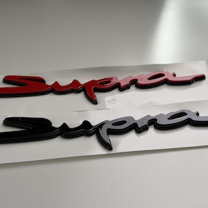 May include: Two adhesive emblems featuring the word Supra in a stylized cursive script. One emblem is glossy red with a black outline, and the other is solid glossy black. Both emblems are mounted on white rectangular backing paper strips.