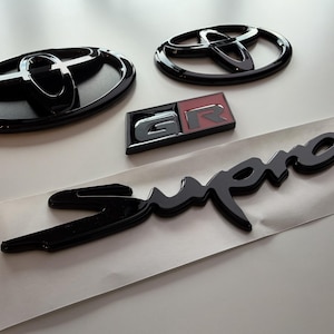 Peut inclure: Un ensemble d'emblèmes automobiles comprenant deux logos ovales Toyota noir brillant, un badge rectangulaire GR avec des accents rouges et argentés, et un emblème Supra en écriture noire fixé sur un support adhésif blanc pour véhicule.