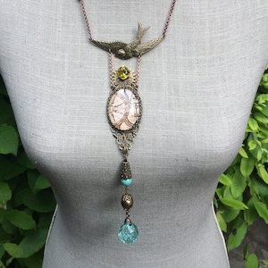May include: A vintage-style necklace with a bronze-colored chain and pendant. The pendant features a bird, a clock face design, and various beads in shades of turquoise, gold, and a clear blue. The necklace is displayed on a mannequin.