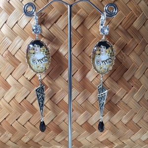 Peut inclure: Une paire de boucles d'oreilles en argent avec une pierre noire en forme de larme suspendue à chaque boucle. Les boucles d'oreilles présentent un pendentif ovale en argent avec un portrait d'une femme en robe dorée. Les boucles d'oreilles sont exposées sur un support en argent.