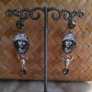 Peut inclure: Une paire de boucles d'oreilles en argent avec des accents de cristal noir et une image ronde d'une personne portant un chapeau. Les boucles d'oreilles sont suspendues à un support en argent.