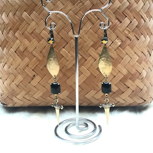 Könnte beinhalten: Ein Paar goldene und schwarze Ohrringe mit einem Dolch-Anhänger. Die Ohrringe haben einen tropfenförmigen goldenen Anhänger mit einem schwarzen quadratischen Stein und einem goldenen Dolch-Anhänger.