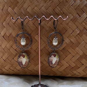Peut inclure: Une paire de boucles d'oreilles de couleur cuivre au design d'inspiration vintage. Chaque boucle d'oreille présente un pendentif circulaire avec une goutte de perle et une image de femme de style camée.