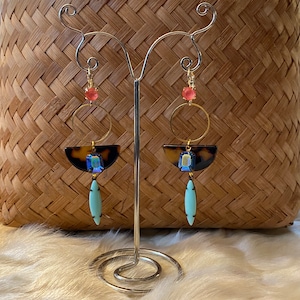 Puede incluir: Un par de pendientes colgantes con un aro dorado, un semicírculo de carey, una piedra preciosa azul y una lágrima turquesa. Los pendientes están colgados de un soporte de plata.