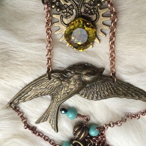 May include: A vintage-style necklace featuring a bronze-colored bird pendant with outstretched wings. The necklace includes a large, faceted yellow-green gemstone, decorative metalwork, and copper-colored chain accents. Small turquoise beads add a pop of color.